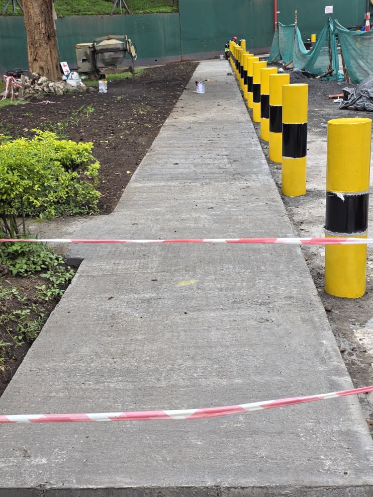 Freshly poured concrete footpath lined with yellow and black safety bollards through landscaped grounds