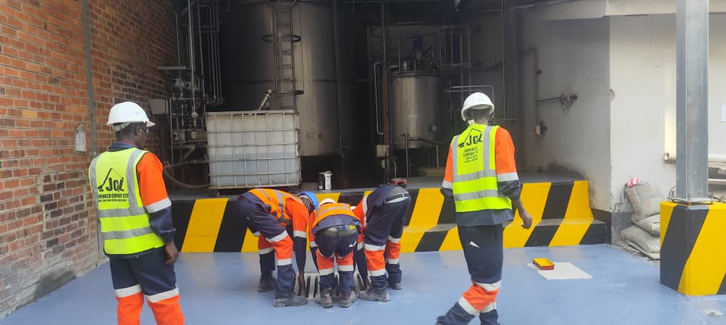 JCL crew inspecting drainage works inside an industrial plant with storage tanks behind