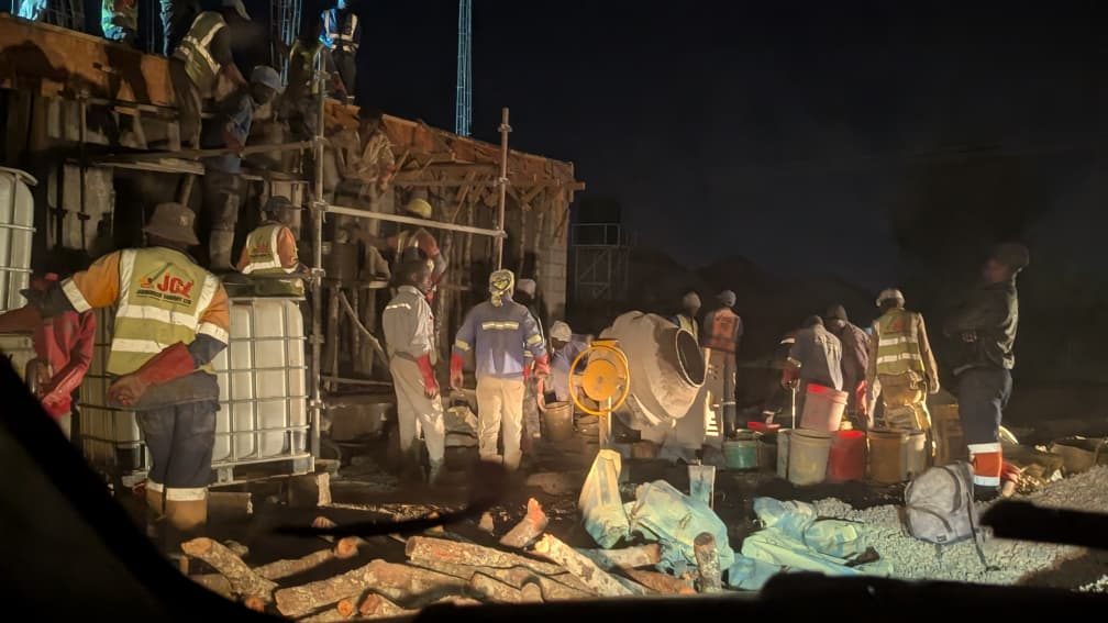 Night shift crew pouring concrete by floodlight around mixers and scaffolding