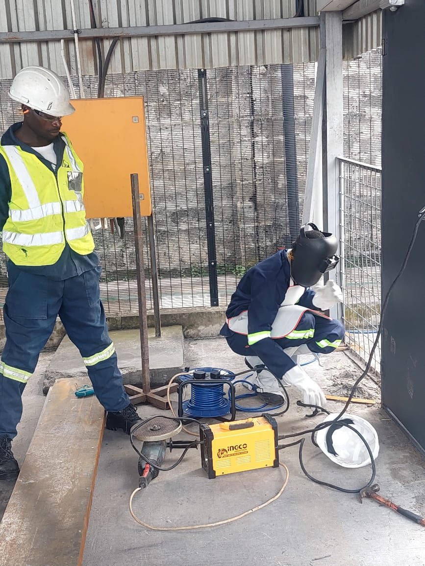 JCL welder in mask fabricating steelwork while supervisor in hi-vis inspects